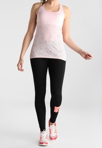 Jasnoróżowy top na ramiączkach z delikatnym wzorem, zestawiony z czarnymi legginsami z różowym akcentem oraz różowo-białymi butami sportowymi.