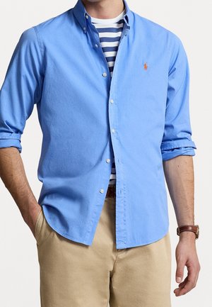 Mężczyzna noszący jasnoniebieską koszulę na guziki na granatowo-białej paskowanej koszulce, beżowe spodnie i brązowy zegarek na nadgarstek.