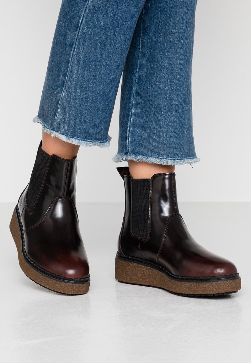 Czarne i burgundowe połyskujące botki do kostki z elastycznymi panelami bocznymi, zaokrąglonymi noskami i jasnobrązowymi fakturowanymi podeszwami na koturnie. Widoczny postrzępiony dół dżinsowy.