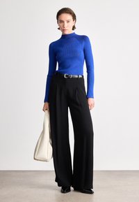 Suéter de gola alta azul com nervuras ajustado, combinado com calças largas pretas e uma mala de creme, apresentando uma textura suave e linhas limpas.