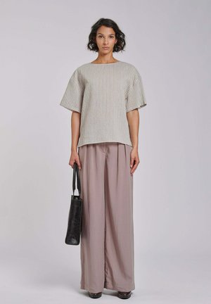 Vrouw met kort krullend haar die een loszittende beige gestreepte top en wijd uitlopende mauve broek draagt, met een zwarte handtas naast zich.