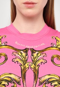 T-shirt rosa con stampa in stile barocco dorato. Presenta un collo rotondo e una texture morbida. Marca "Versace Jeans Couture" stampata sul fronte.