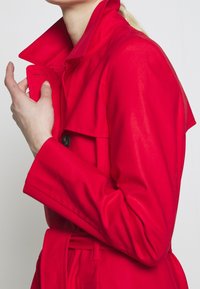 Trench-coat rouge avec un col montant, une fermeture à boutons et une ceinture nouée. Texture lisse avec des détails de couture visibles et des manches surdimensionnées.
