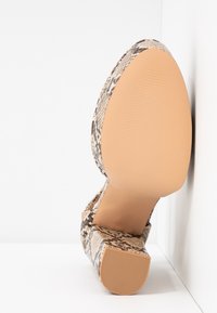 Chaussure à talon épais unique avec semelle beige et dessus à motif peau de serpent, positionnée semelle vers le haut contre un fond blanc d'angle.