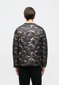 Rövid hajú személy kék és bézs absztrakt mintázatú steppelt dzsekit visel, háttal áll egyszerű fehér háttér előtt.