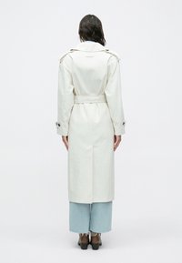 Hvid trenchcoat lavet af letvægtsstof, med bælte i taljen, skulderepauletter og lange ærmer. Bæres over brede blå jeans.