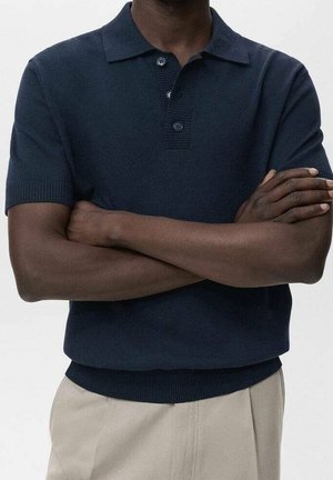 Mann mit dunkler Haut, der ein marineblaues kurzärmeliges Poloshirt und beige Hosen trägt, steht mit verschränkten Armen vor der Brust.