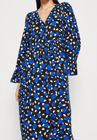 Abito nero con pois astratti blu e bianchi, scollatura a V, maniche lunghe con polsini arricciati e design con vita arricciata.