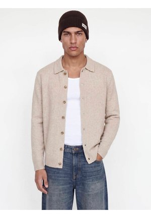 Jeune homme portant un cardigan beige à boutons, un t-shirt blanc, un jean bleu et un bonnet tricoté marron foncé sur fond blanc.