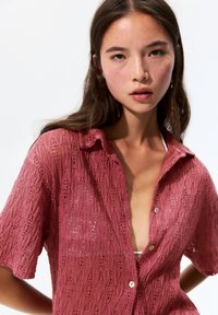 Gebreide, kortslippige shirt in stoffige rozenkleur met een kanten patroon, voorzien van een kraag en knoopsluiting, waarbij een witte onderkleding zichtbaar is.