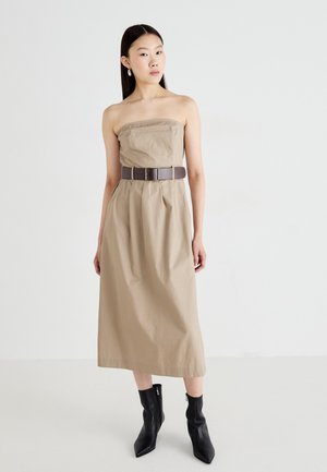 Vestido beige sin mangas con un torso ajustado y una cintura ceñida, combinado con un cinturón marrón. La modelo lleva botas negras hasta el tobillo. Textura simple.