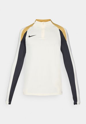 Maglietta sportiva a maniche lunghe color crema con dettagli neri e dorati. Presenta un colletto con mezza zip e un design aderente per l'uso atletico.
