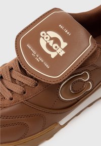 Bruine Coach sneaker met gevlochten veters en een reliëflogo op de tong en zijkant, voorzien van de teksten "est. 1941" en "Ontworpen in New York, NY, VS".