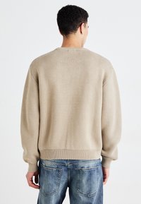 Beige strikket sweater med en afslappet pasform, ribbede manchetter og kant; båret over blå denim jeans. Viser et struktureret, vævet mønster.