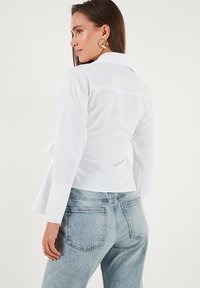 Woman wearing a white long-sleeve shirt and light blue jeans, facing away, with gold hoop earrings and long brown hair.