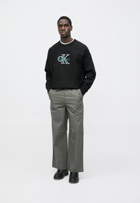 Black crewneck sweatshirt with "CK" logo, paired with light green wide-leg trousers. Black shoes complete the outfit. Minimalist design.