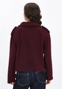 Burgunderfarbene, taillierte Jacke mit Schulterklappen, langen Ärmeln, weicher Textur und rundem Kragen. Kombiniert mit dunkelblauen Jeans.