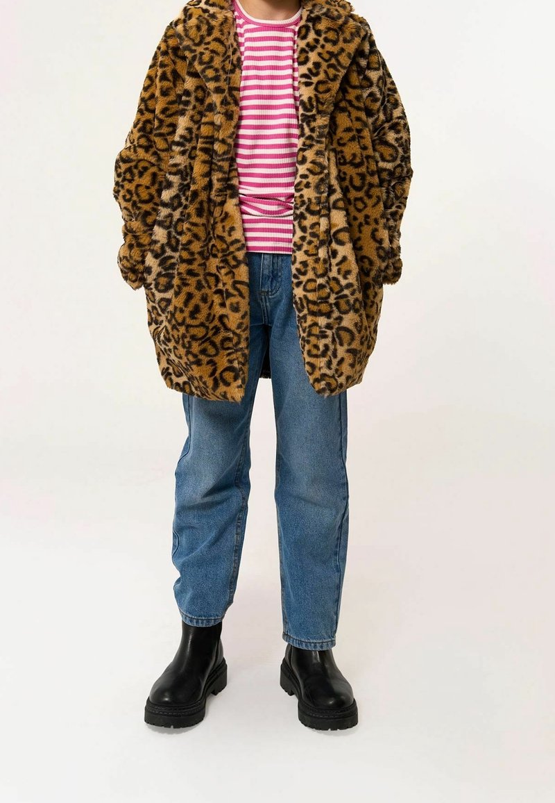 Personne portant un manteau en fausse fourrure imprimé léopard, une chemise rayée rose et blanche, un jean bleu et des bottines noires, debout sur un fond uni.