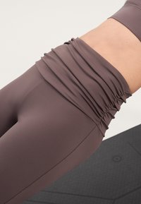 Barna, katecsoportos sport leggings, ráncolt derékkal, sima, rugalmas anyagból. Textúrázott fekete matrac a háttérben.