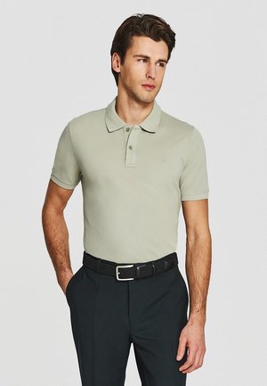 Mężczyzna w jasnobeżowej koszulce polo wpuszczonej w ciemne spodnie z czarnym plecionym paskiem, stojący z jedną ręką w kieszeni.