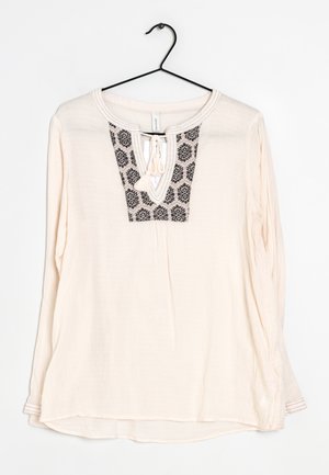 Blouse crème à manches longues avec broderie géométrique noire sur la poitrine et détail de lien, suspendue sur un cintre noir sur fond blanc.