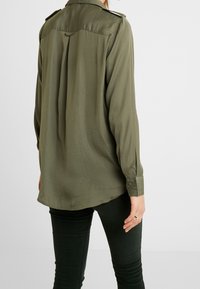 Camicia verde oliva con maniche lunghe, polsini abbottonati e una piega sul retro. Tessuto morbido, vestibilità aderente e orlo arrotondato. Abbinata a pantaloni scuri.