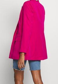 Fuchsiové sako s přizpůsobeným střihem, dvěma předními knoflíky a dlouhými rukávy. Nošené s roztrhanými denimovými šortkami pro vzhled s kombinací různých textur.