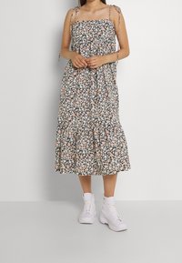 Blumenkleid in Midi-Länge mit dünnen Bindebändern, schwarzer Hintergrund geschmückt mit pastellfarbenen Blumen, gerafftem Oberteil und fransigem Saum. Getragen mit weißen Sneakers.