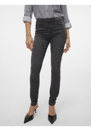 Femme portant un jean gris foncé ajusté, des talons noirs à bout pointu, et une chemise rayée à boutons avec les manches retroussées, debout devant un fond uni.