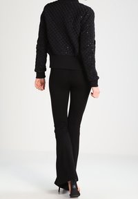 Czarna pikowana kurtka bomber z prążkowanym dołem i mankietami, w zestawieniu z czarnymi spodniami w dzwony i butami na obcasie. Gładka faktura całości.