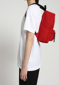 Roter Stoffrucksack mit rechteckiger Form, schwarzen verstellbaren Trägern und einer Vordertasche, getragen über einem weißen Hemd und dunklen Hosen.