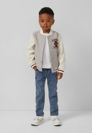 Grijs en crèmekleurig varsityjack met geborduurde tekst, wit T-shirt, blauwe jeans en witte sneakers. Gebreide boorden en drukknoopsluiting.