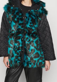 Faux fur-väst med svarta quiltade ärmar, med ett mönster av turkos och svart leopard, knäppning med knappar och en huva.