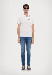 Biele polo tričko s čiernymi akcentmi a logom EA7, kombinované s modrými úzkymi džínsami a tmavými teniskami, nosené stojacim mužským modelom.