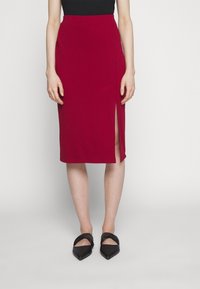 Femme portant une jupe rouge mi-longue avec une fente à l'avant, un haut noir et des ballerines pointues noires avec des brides torsadées, debout devant un fond uni.