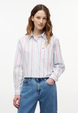 Νεαρή γυναίκα με κυματιστά μαλλιά, που φοράει ένα λευκό πουκάμισο με κόκκινες κάθετες ρίγες και μπλε τζιν, στέκεται με το ένα χέρι στην τσέπη.