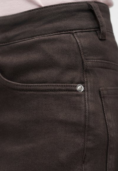 Κοντινό πλάνο τσέπης σκούρου καφέ τζιν παντελονιού με ορατή ραφή, καρφίτσα και ζώνη μέσης, σε φόντο ανοιχτού υφάσματος.