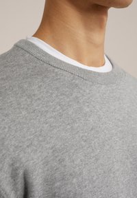 Suéter gris con una tela texturizada, que presenta un cuello redondo y un collar de punto acanalado. Llevado sobre una camiseta blanca por debajo.