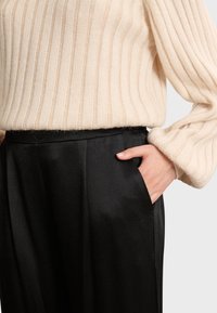 Bežani rebrasti pulover s dugim rukavima uparen s crnim satenskim hlačama visokog struka, koje imaju bočnu džep i glatku teksturu.