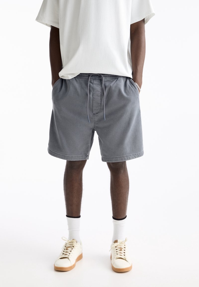Szare szorty bawełniane z elastycznym paskiem i sznurkiem do regulacji, z bocznymi kieszeniami i luźnym krojem, w połączeniu z białymi trampkami i skarpetkami.