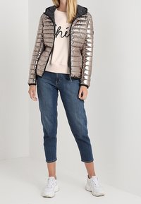 Giacca puffer argentata con cappuccio nero, chiusura con zip, abbinata a un maglione rosa chiaro e jeans blu cropped. Sneakers bianche.