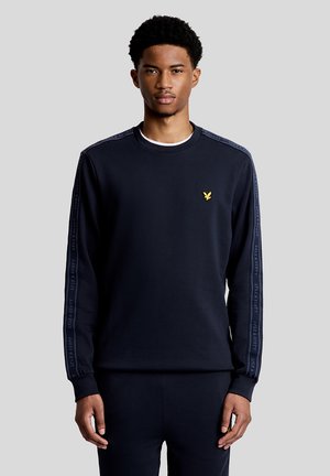 Jonge man die een zwarte trui draagt met een logo op de borst en merkstrepen langs beide mouwen, staand tegen een effen grijze achtergrond.