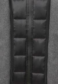 Zwart gewatteerd stof met een doorgestikt patroon, met een centrale ritssluiting, geplaatst tegen een lichtgrijze gestructureerde achtergrond.