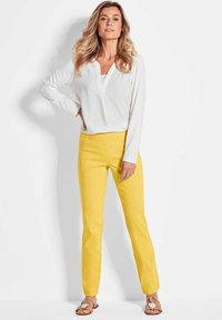 Witte blouse met lange mouwen en V-hals, gecombineerd met strakke gele broek. De look wordt compleet gemaakt met platte sandalen met ronde accenten.