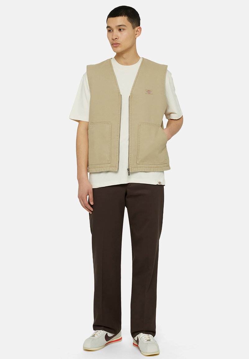 Beige Utility-Weste mit zwei vorderen Taschen, getragen über einem weißen T-Shirt, kombiniert mit braunen Hosen und grauen Turnschuhen. Neutrale Farbpalette.