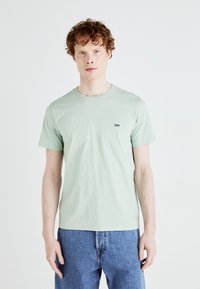 Camiseta de algodón verde claro con cuello redondo. Presenta un pequeño logo negro de "Lee" en el pecho. El modelo lleva pantalones vaqueros de mezclilla azul.