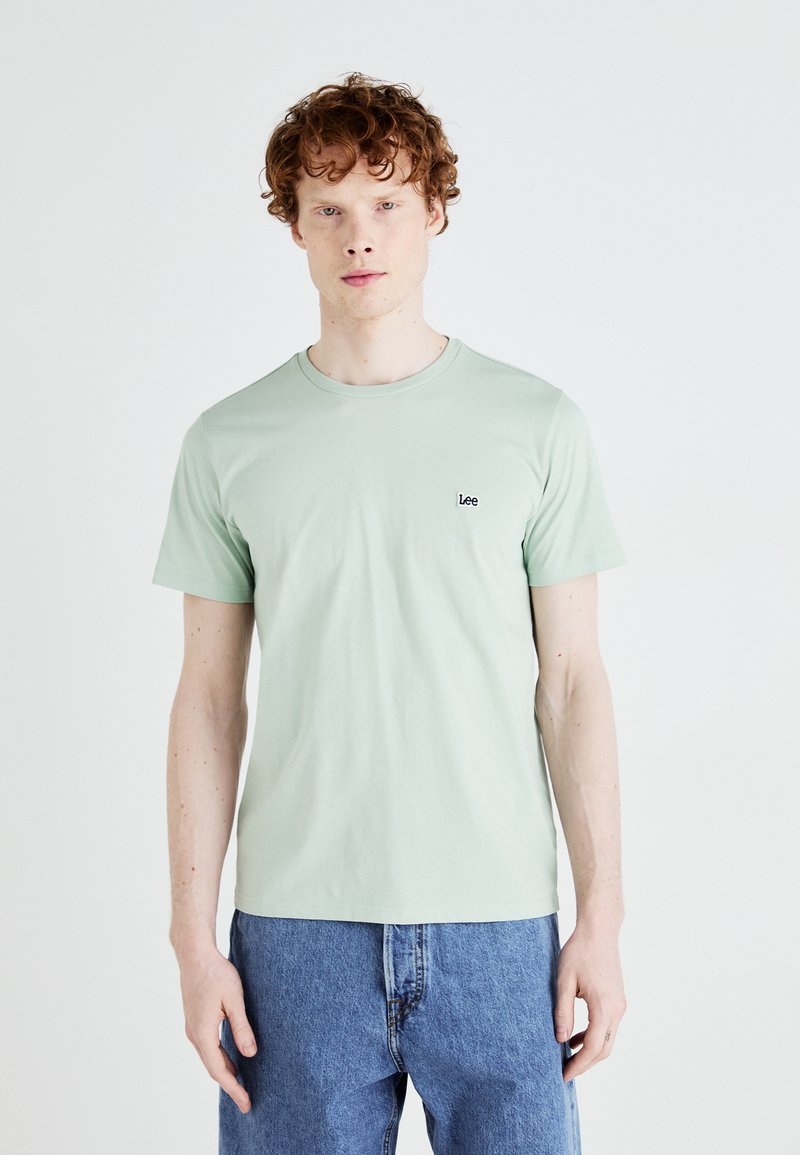 Camiseta de algodón verde claro con cuello redondo. Presenta un pequeño logo negro de "Lee" en el pecho. El modelo lleva pantalones vaqueros de mezclilla azul.