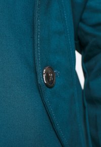 Turkusowa kurtka o gładkiej fakturze, z pojedynczym dużym guzikiem z wygrawerowanym napisem "BEN SHERMAN" i widocznymi szwami na krawędziach.