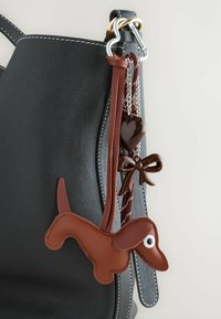 Sort lædertaske med hvid syning, udstyret med en charm i brunt læder formet som en dachs, et mørkt hjerte og en brun sløjfe akcent på en dekorativ kæde.