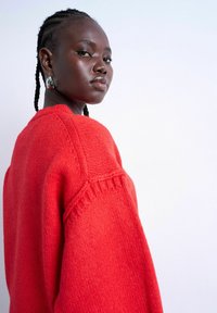 Femme aux cheveux tressés portant un pull en maille rouge vif et des boucles d'oreilles en pierre verte, regardant par-dessus son épaule sur un fond clair uni.